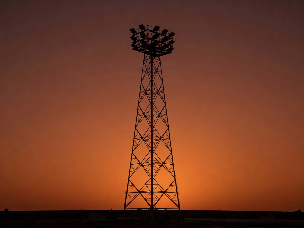 A tall lattice floodlight pylon silhouetted against a dark orange dusk sky